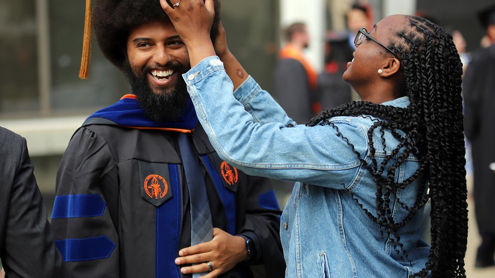 A woman with long dark braids and a denim jacket straightens the mortarboard of a smiling man who is graduating from Caltech