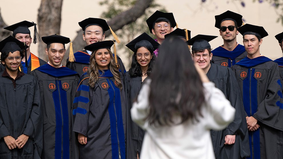 A woman in a white jacket with long dark hair takes a photograph of 12 Caltech graduates