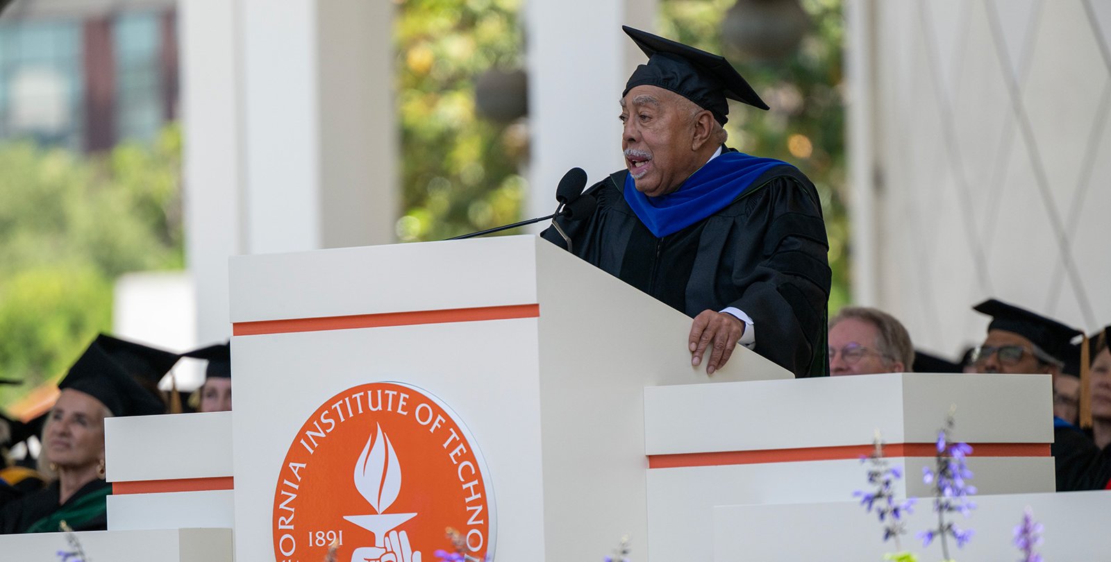 An older man wearing graduation robes with a blue stole speaks to an audience from a lecturn