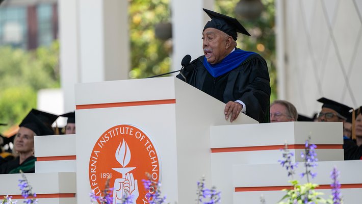 An older man wearing graduation robes with a blue stole speaks to an audience from a lecturn