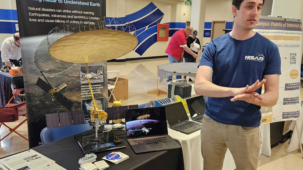 A person speaks in front of an exhibit of materials related to the NISAR mission