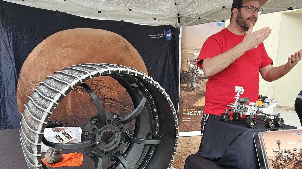 A person speaks in front of an exhibit of materials about Mars exploration missions, a rover wheel in the foreground