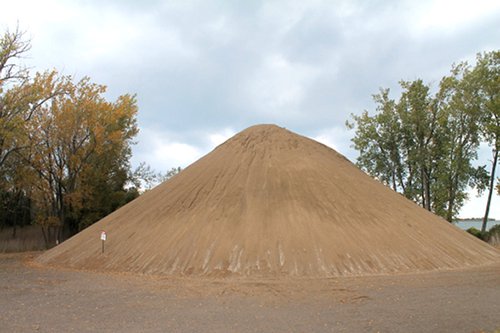 A large pile of sand sits in a vacant lot with trees behind it.