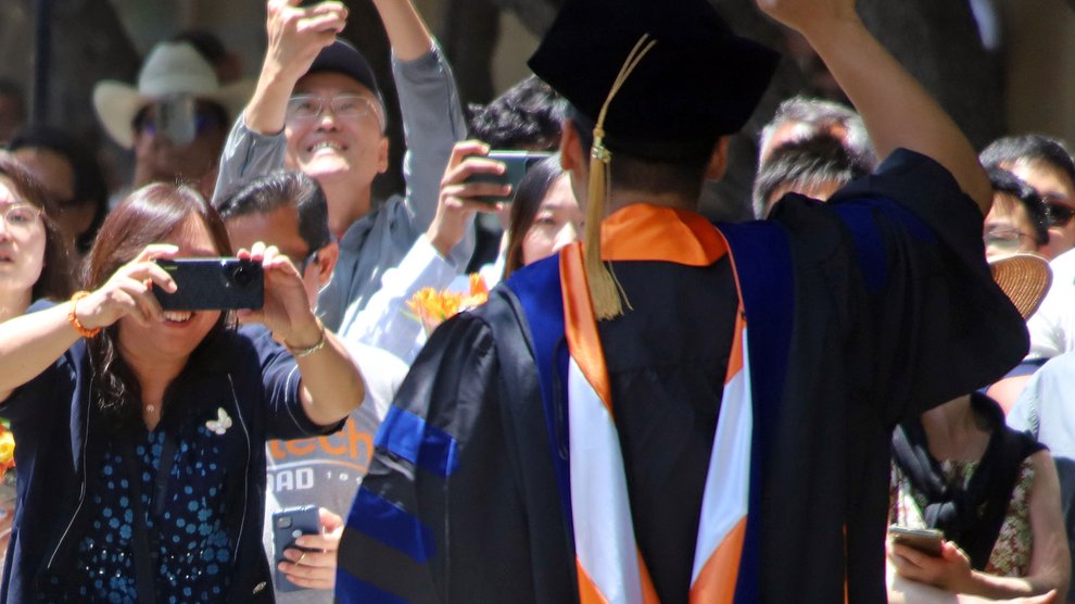 A man standing with his back to the camera wearing graduation robes waves to a group of people who are photographing him