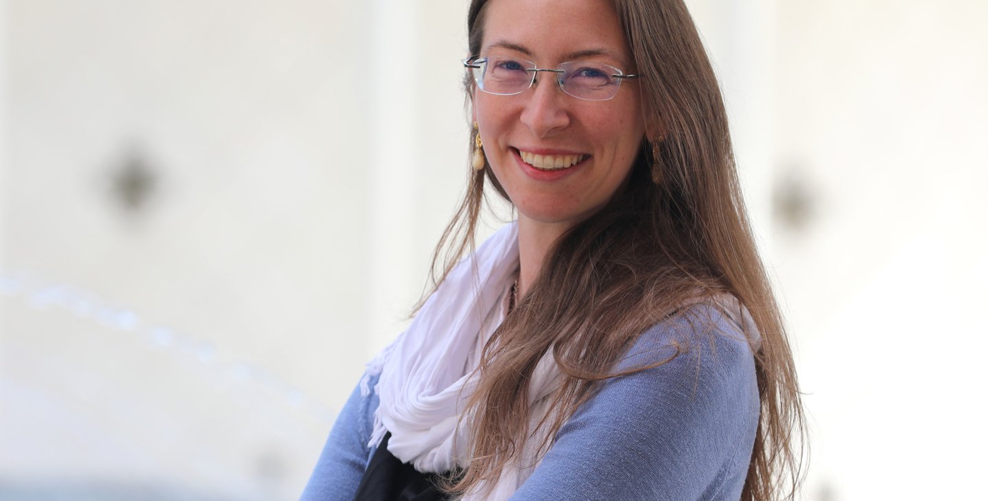 Franca Hoffmann, with long light brown hair and glasses, wearing a white scarf and a light blue sweater, stands outside Beckman Auditorium on the Caltech campus.