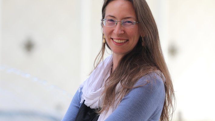 Franca Hoffmann, with long light brown hair and glasses, wearing a white scarf and a light blue sweater, stands outside Beckman Auditorium on the Caltech campus.