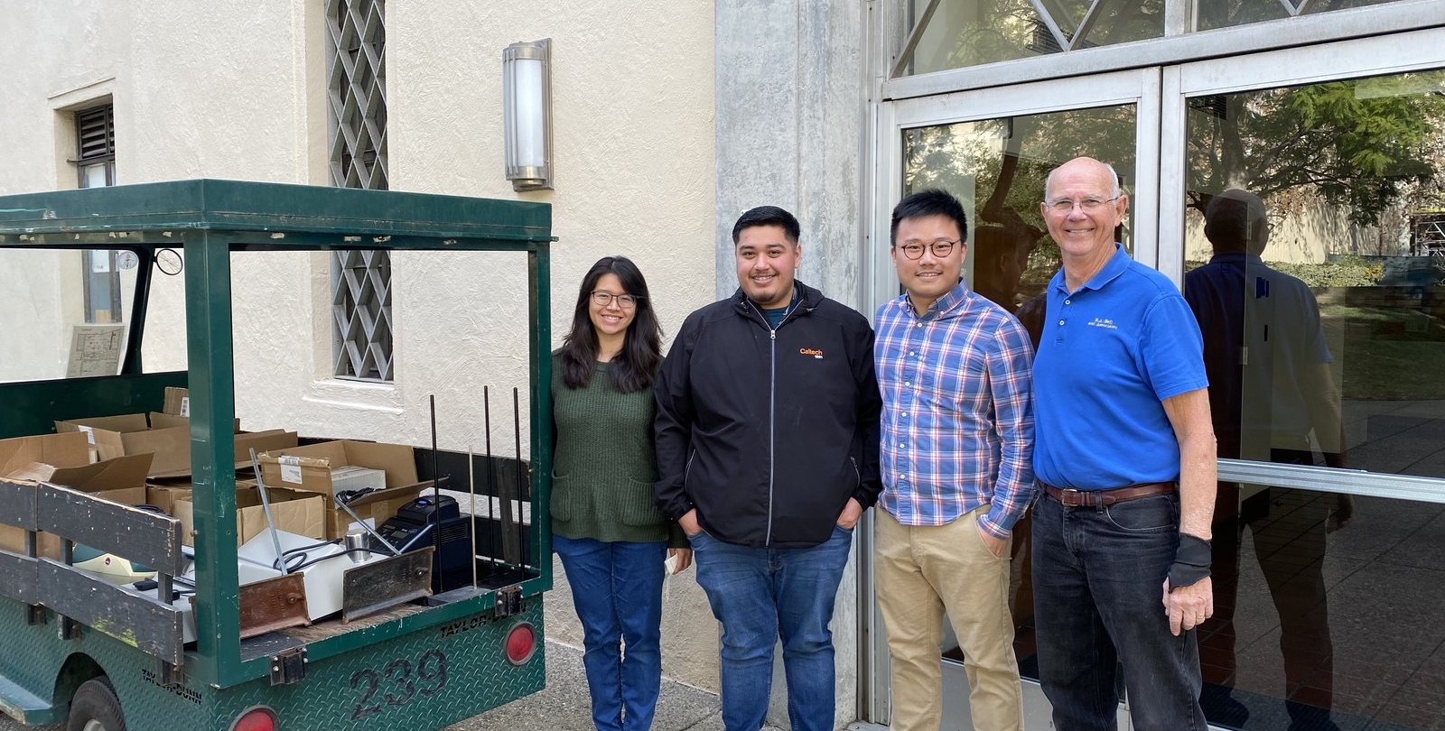 Four people stand smiling near an electric cart loaded with equipment in boxes, a building in the background