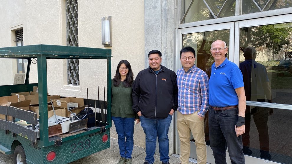 Four people stand smiling near an electric cart loaded with equipment in boxes, a building in the background