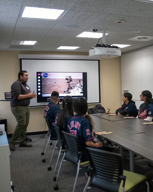 A standing person talks in front of a slide as a small group of people seated at a table watches