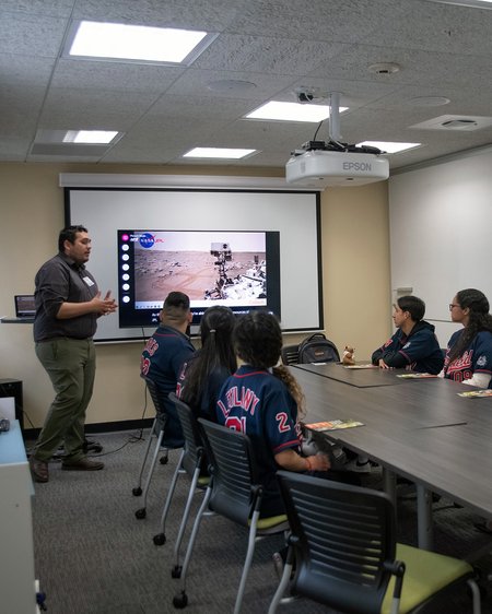 A standing person talks in front of a slide as a small group of people seated at a table watches
