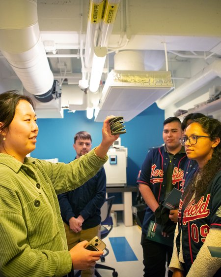 In a laboratory setting, as four people look on, another holds up a green metallic object like a spiral cage with a solid lid and floor and a ball inside it.