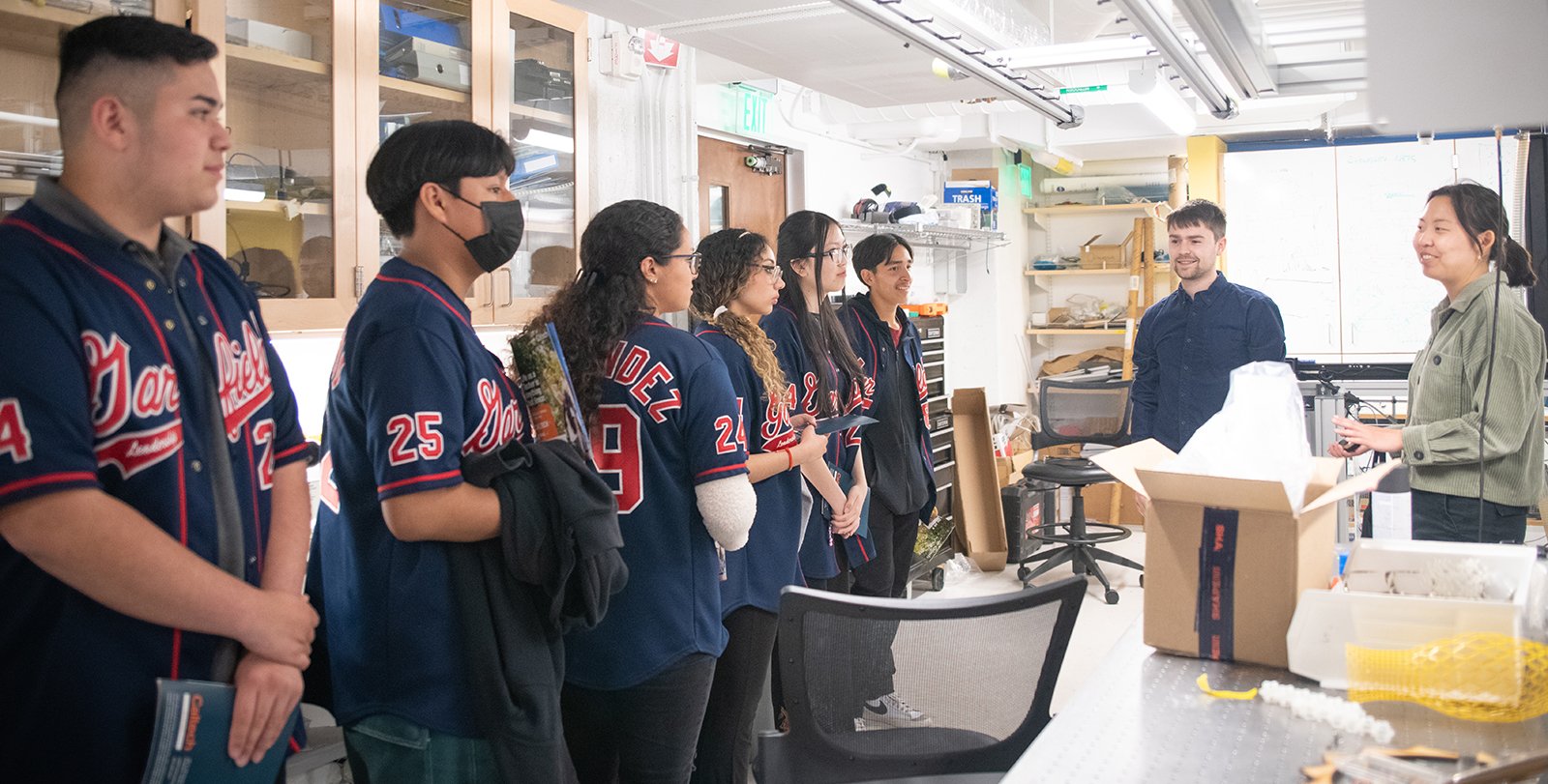 In a laboratory, six people in Garfield High School jackets watch two people who are facing them, smiling.