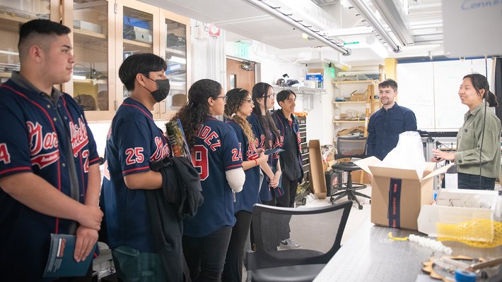 In a laboratory, six people in Garfield High School jackets watch two people who are facing them, smiling.