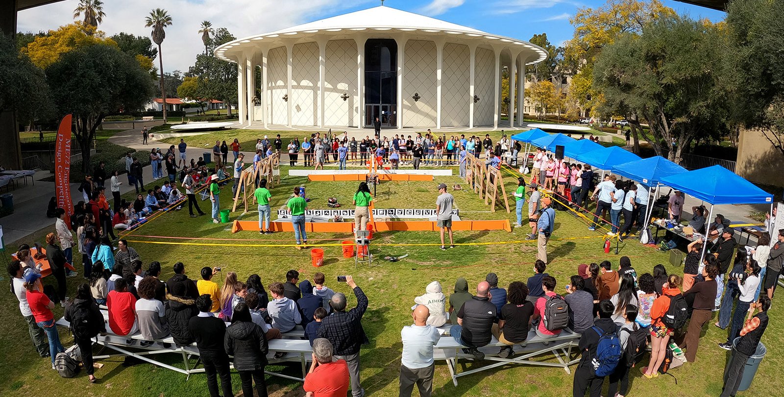A crowd of people watch the ME72 competition in front of the Beckman Auditorium, a round white building with columns