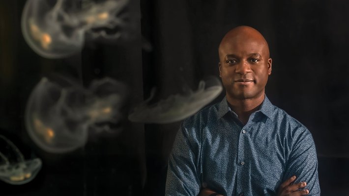 Photo of John Dabiri wearing denim shirt, posed in front black background featuring jellyfish