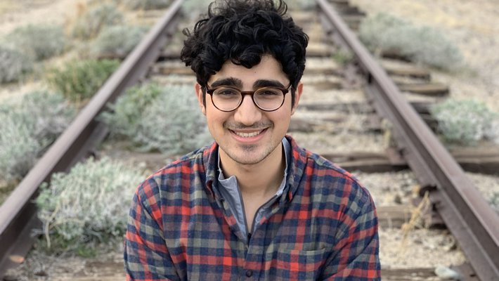 Portrait of Booeshagi sitting outdoors on train tracks, smiling at the camera, wearing round glasses and a plaid shirt