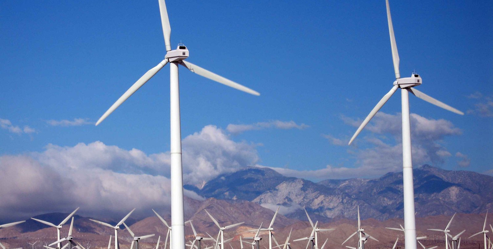 Wind turbines spin in front of a backdrop of clouds and mountains.