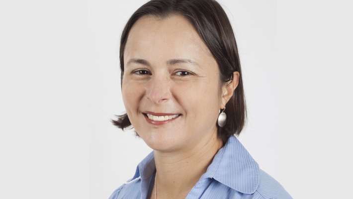 Headshot of alumna Gypsy Achong against a white background. She has dark, shoulder length hair and is wearing a blue shirt.