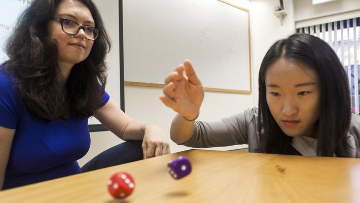 A woman in a blue shirt looks on as a student rolls a pair of dice onto a table