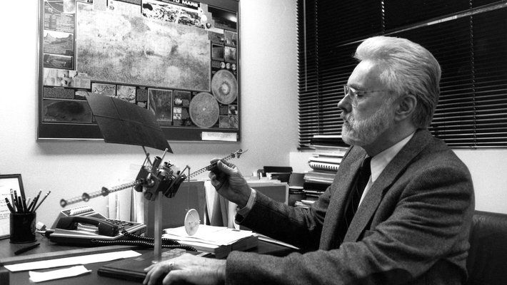 Arden Albee sits at a desk wearing a tie and sport jacket, looking at a model spacecraft for a Mars mission. There is a large poster on the wall titled "An Explorer's Guide to Mars."