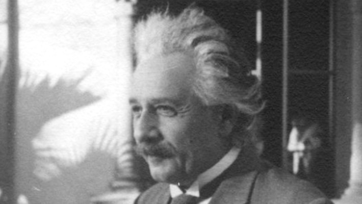 A black and white photograph of Albert Einstein. He has silver hair brushed back from his face and a dark mustache. He wears a three-piece suit and tie.