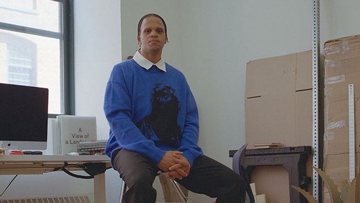 A man in a blue sweater, black pants, and black shoes sits with his hands in his lap looking into the camera. He is seated on a stool in an art studio, surrounded by cardboard, elements of a sculpture, and a desk with a computer and books.