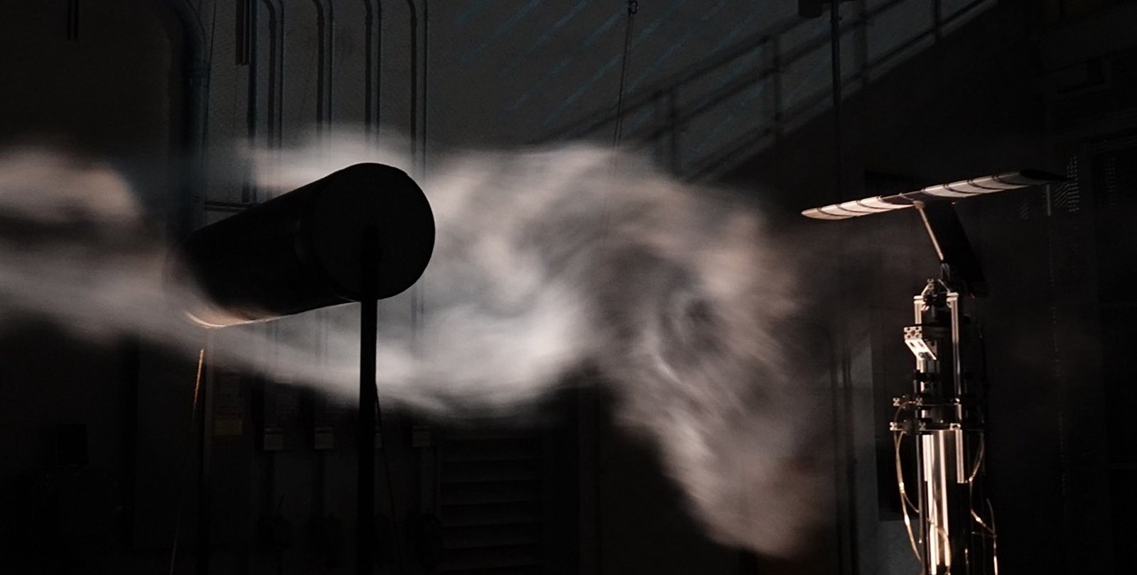 The turbulent wake of a cylinder can be seen in illuminated smoke in a darkened wind tunnel. An airfoil instrument is set up on the right side of the image. A large cylinder is on the left side of the image, creating turbulence.