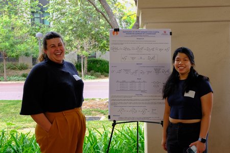 Two young women are shown standing on either side of a chemistry poster with greenery in the background.