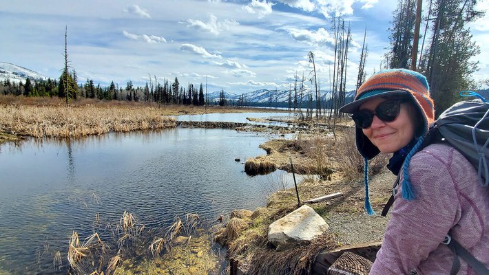 Woman in winter hat, sunglasses, and backpack in front of a body of water, leafless trees and a cloud-speckled blue sky