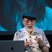 Kip Thorne, in a beret and button-down shirt, gestures as he speaks near a podium