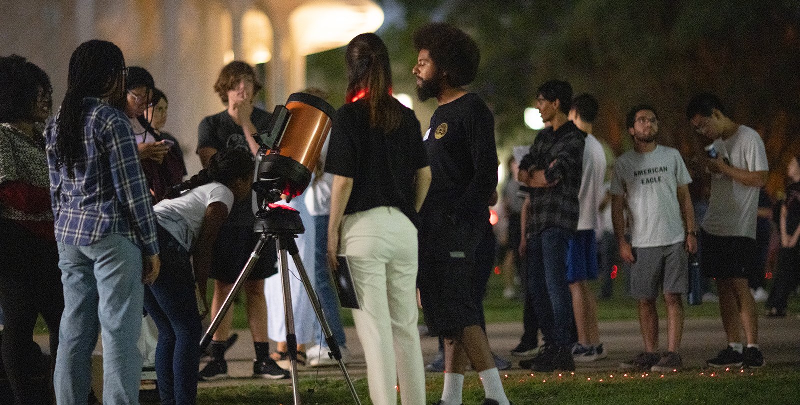People gather around a telescope on a lawn with Beckman Auditorium in the background