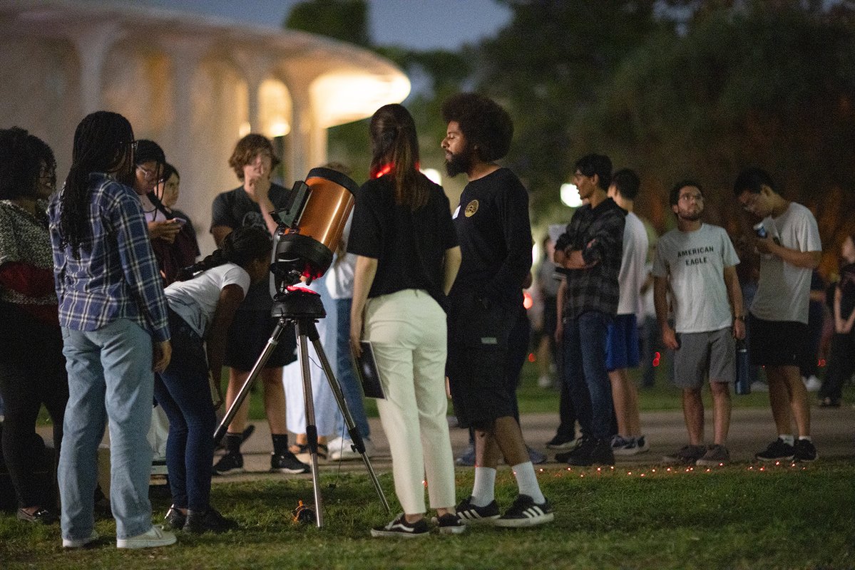 People gather around a telescope on a lawn with Beckman Auditorium in the background