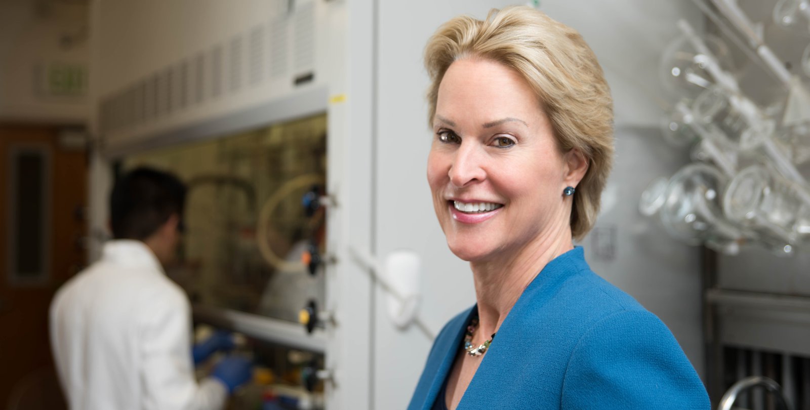 A portrait of Frances Arnold in her laboratory. She wears a blue blazer and smiles at the camera. In the background are laboratory glassware and a researcher at work.
