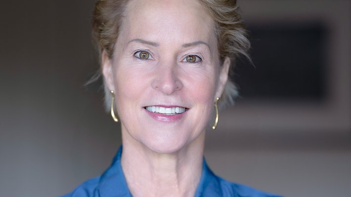Head shot of a woman with short blond hair wearing a blue collared shirt and gold earrings.