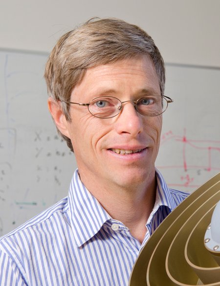 A portrait of Jean-Philippe Avouac smiling . He is shown in front of a white board and with a model.