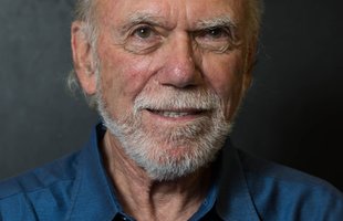 Portrait of Barry Barish. He wears a navy blue shirt and has a white mustache and beard.