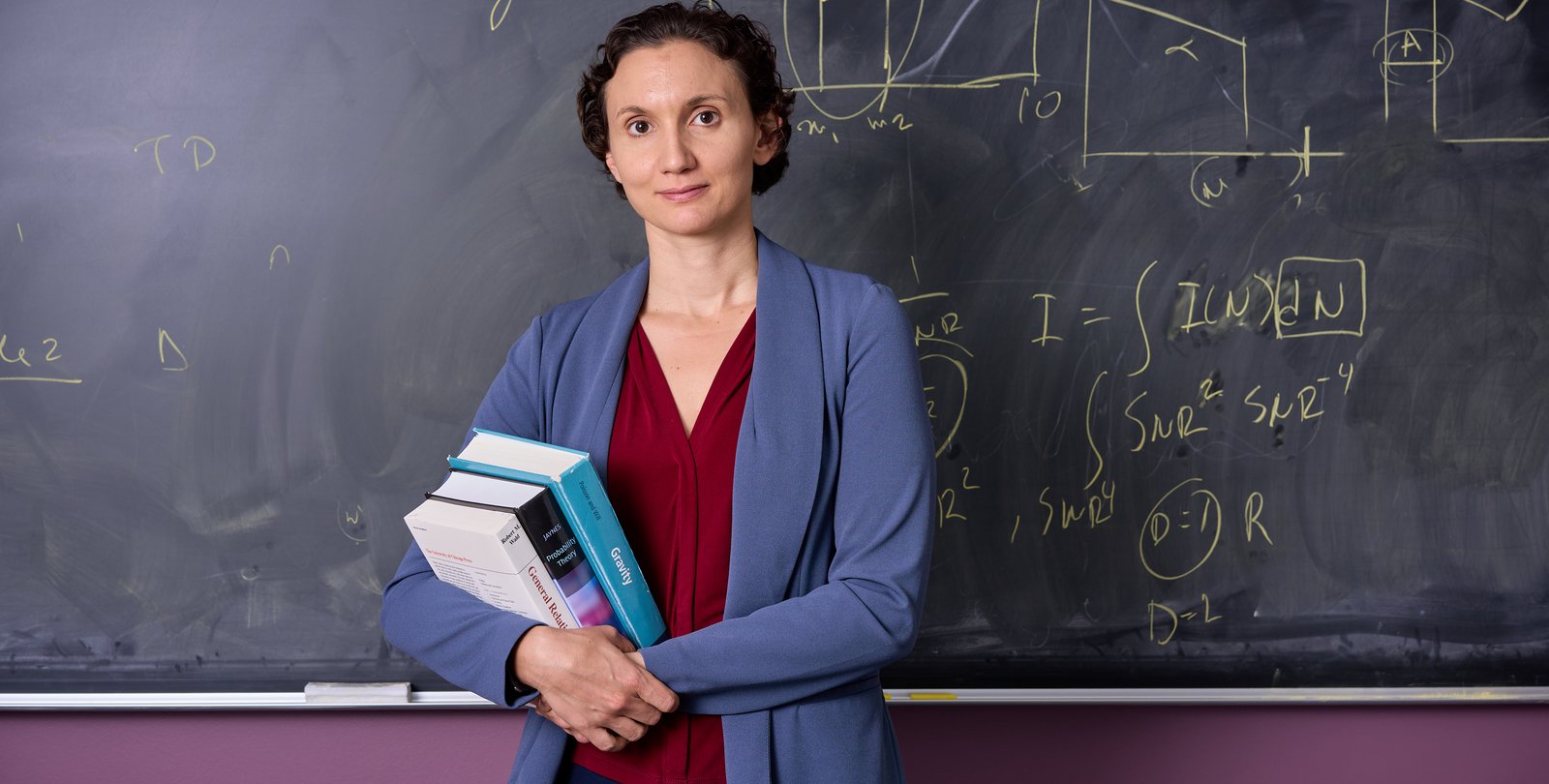 Image of Katerina Chatziioannou standing in front of chalk board holding books in grey blazer and burgandy blouse