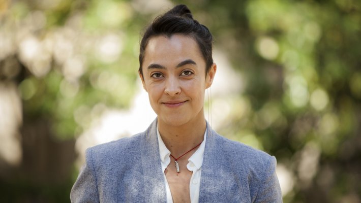 A portrait of Maura Dykstra. She wears a blazer and appears in front of plants outdoors.