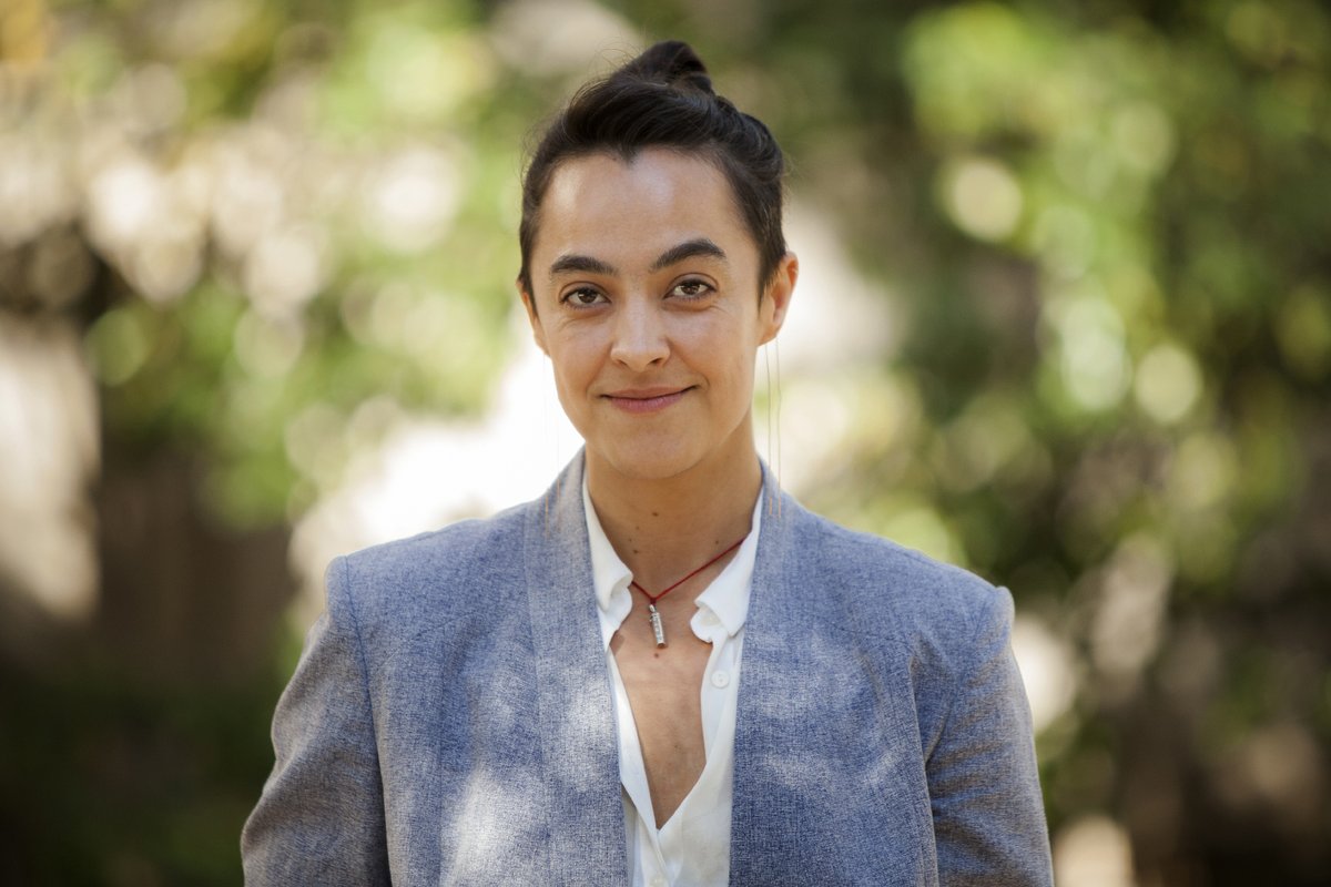 A portrait of Maura Dykstra. She wears a blazer and appears in front of plants outdoors.