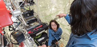 Graduate student Stephanie Kong wearing a blue lab coat looks up at a colleague while adjusting equipment inside a room that holds an atmospheric chamber. She is surrounded by wires, equipment, and the floor to ceiling plastic of the chamber walls