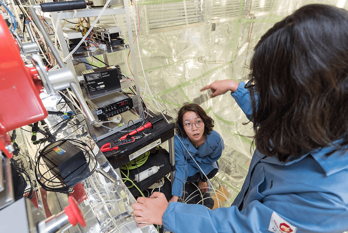 Graduate student Stephanie Kong wearing a blue lab coat looks up at a colleague while adjusting equipment inside a room that holds an atmospheric chamber. She is surrounded by wires, equipment, and the floor to ceiling plastic of the chamber walls