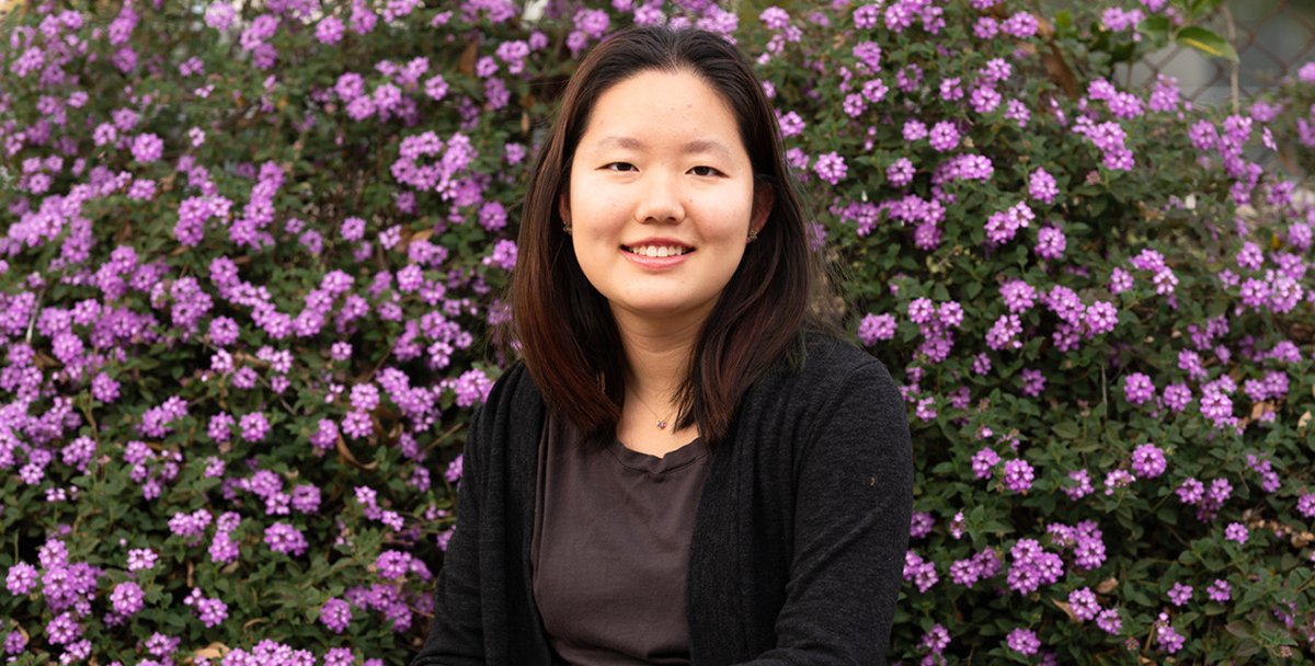 Yiyi Cai, in a brown shirt and black sweater, sits in front of a bush covered in purple flowers.