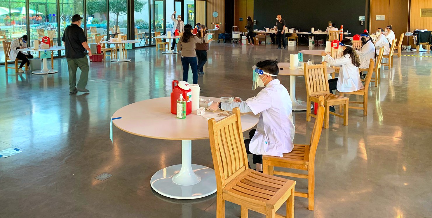 White-coated workers sit at widely spaced tables with vaccines shots at the ready.
