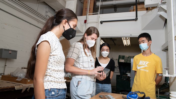 Four people in K95-style masks and short-sleeved shirts stand at a tall table in a narrow, white-painted laboratory room, discussing and look at a gray rock in Bednarick's hands.