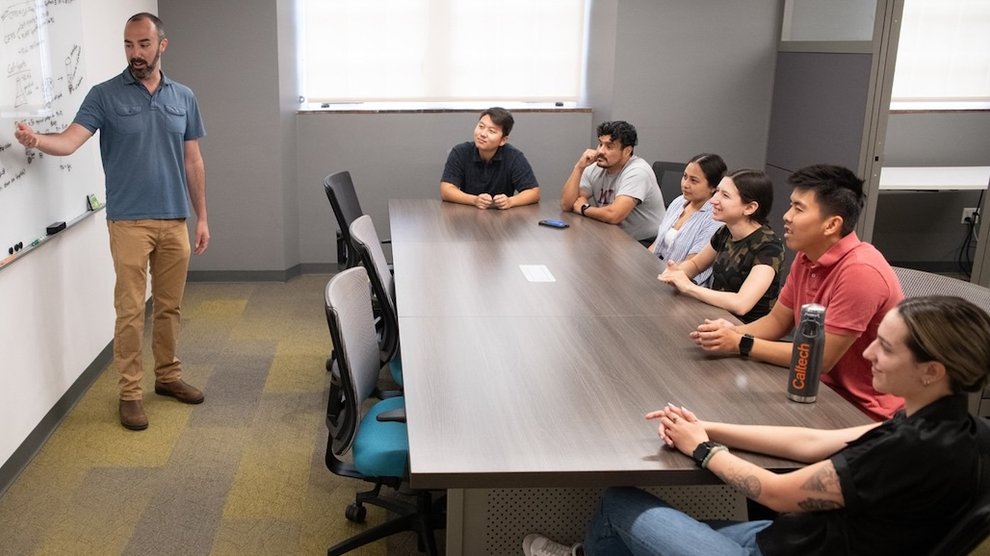 A person at a whiteboard talks to six people seated at a table and listening eagerly