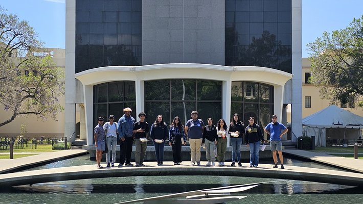 Twelve people stand close together in a line on a bridge over a reflecting pond in front of a building.