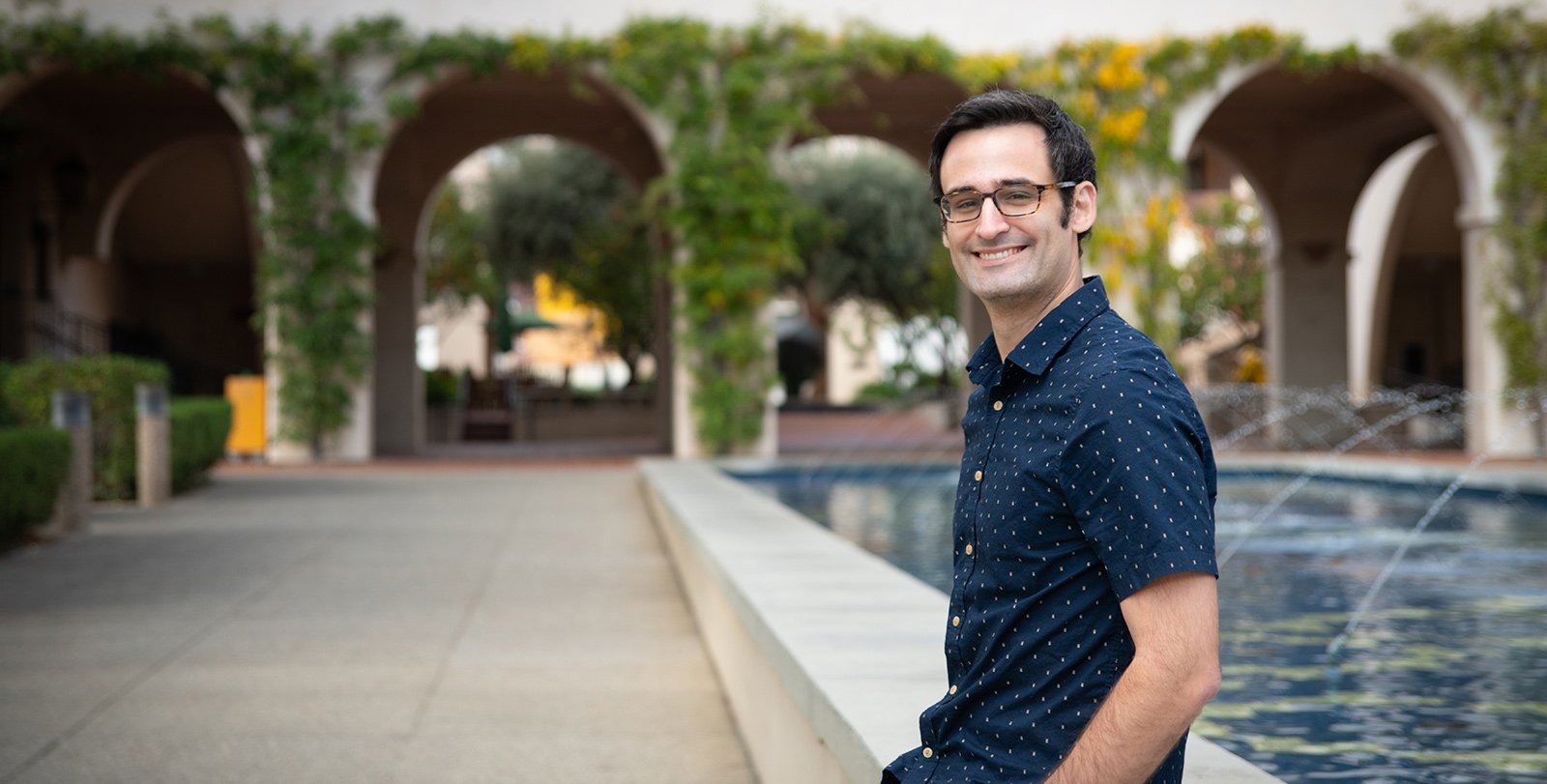 A portrait of Peter Caradonna. He wears a short-sleeve button up shirt and slacks. He sits casually in front of a fountain, smiling.