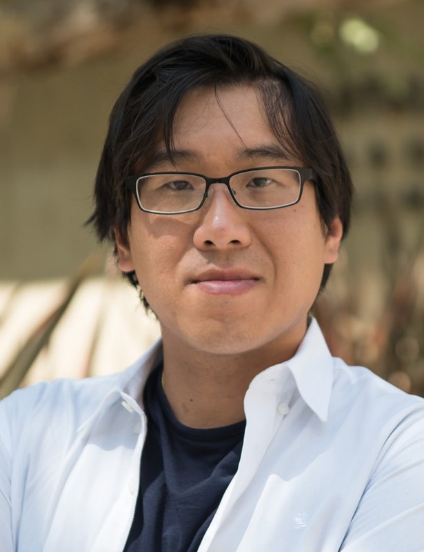 A portrait of Garnet Chan wearing a white collared shirt over a black t-shirt. He has dark-framed glasses.
