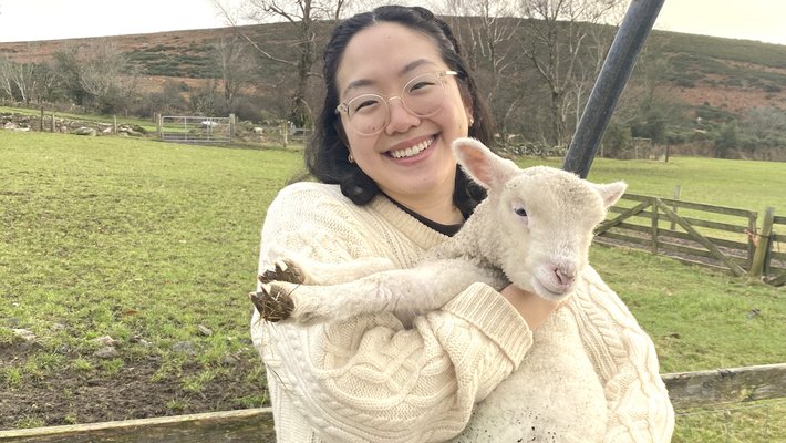 Young woman in a white sweater and black pants holds a sheep.