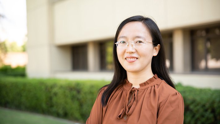 A portrait of Shasha Chong. She smiles at the camera and stands in front of a campus building with trees and grass in the background.